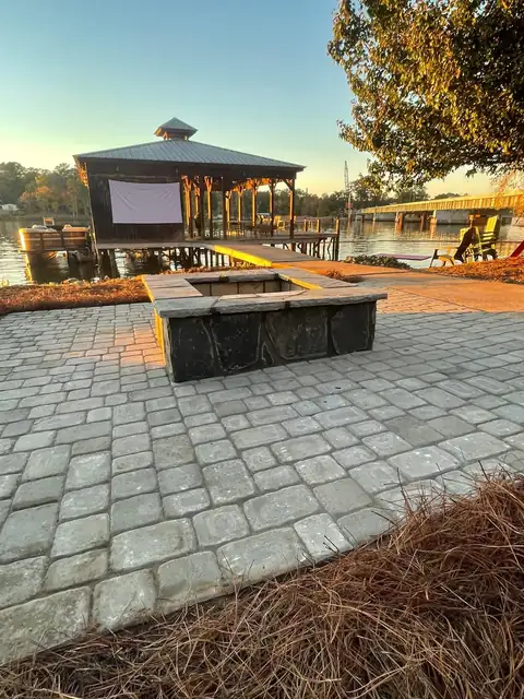 Square stone fire pit on a paver patio at a lake house, boathouse behind it at sunset